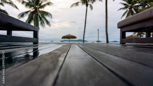 close up of wet wooden floor between wooden lounge chairs after morning rain by the swimming pool, visible thatched roof and several coconut trees. vacation with family at a hotel