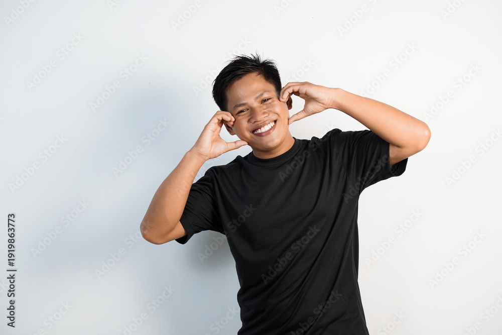 Ταπετσαρία A cheerful young Asian man wearing a black shirt expresses happiness with a heart shaped hand gesture placed on his cheek against a white background
