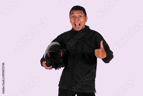 An expressive Indonesian man smiling on an isolated white background while giving a thumbs up (okay symbol) while wearing a jacket carrying a helmet