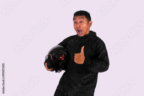 An expressive Indonesian man smiling on an isolated white background while giving a thumbs up (okay symbol) while wearing a jacket carrying a helmet
