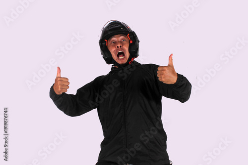 An expressive Indonesian man smiles on an isolated white background while giving a thumbs up (okay symbol) while wearing a helmet and jacket.