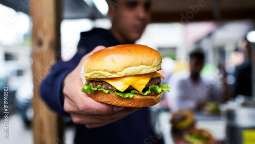 A hand presents a juicy cheeseburger with melted cheese and fresh lettuce in an outdoor setting, against a blurred street background.