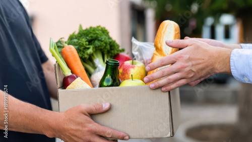 A delivery person hands a brown cardboard box with fresh produce and bread to a customer outdoors, blurring the urban background.