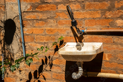 A functional outdoor white plastic utility sink mounted on a rustic red brick wall with exposed plumbing, a flexible metal faucet, and green foliage under bright sunlight with harsh shadows
