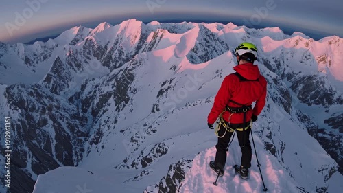 Mountaineer standing on snowy ridge overlooking vast mountain range. Climber on high altitude peak at dawn. Extreme winter adventure