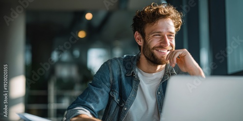Positive young man using laptop computer for video call and smiling while sitting at desk in modern home office space with natural light