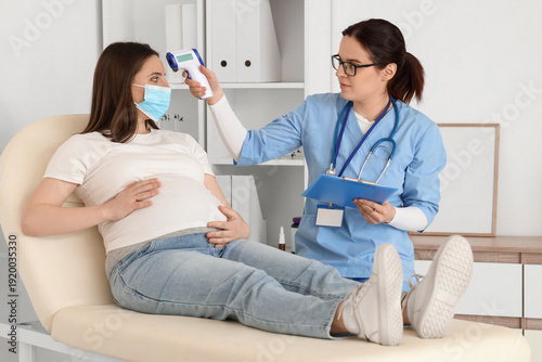 Young pregnant woman in medical mask with doctor measuring temperature at hos...