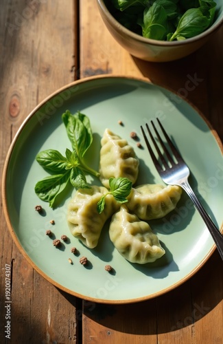 Plate with four green dumplings garnished with basil leaves on wooden table. Fork rests on plate, scattered peppercorns add spice. Natural sunlight creates shadows.