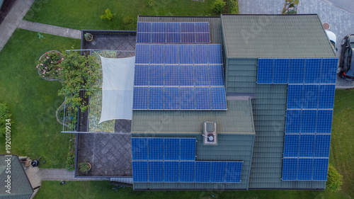 Top-down aerial view of a residential house with multiple solar panels installed on the roof, surrounded by a green garden and yard, showcasing sustainable living and renewable energy in a suburban