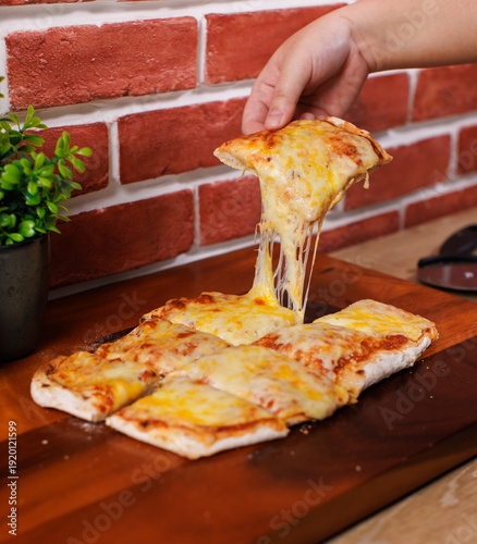 A slice hot traditional pinsa romana pizza square shaped toppings with stretching cheese golden yellow color on wooden cutting board and brick wall tasty snack fresh delicious gourmet italian cuisine.