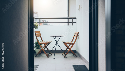 Minimalist apartment balcony seen from inside, featuring simple wooden chairs and a table in a clean, bright outdoor space.
