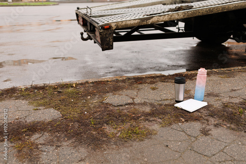 A coffee cup and a water bottle sit on the ground by a truck in a wet city space