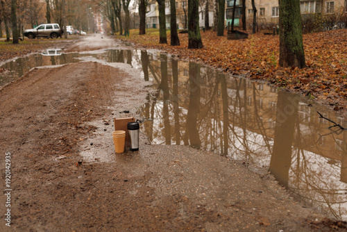 Two coffee cups are on a dirt road next to a puddle, surrounded by wet ground and trees