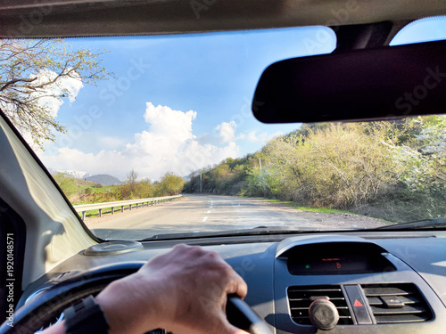 view through car windshield of natural landscape with green field, white clouds in blue sky. Hand of woman on steering wheel of car. Female traveler