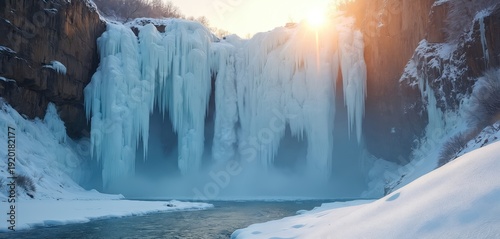 Frozen waterfall cascades down icy cliffs surrounded by snowy landscape. Sunlight shines through mist creating a magical winter scene with icicles and frozen water formations.