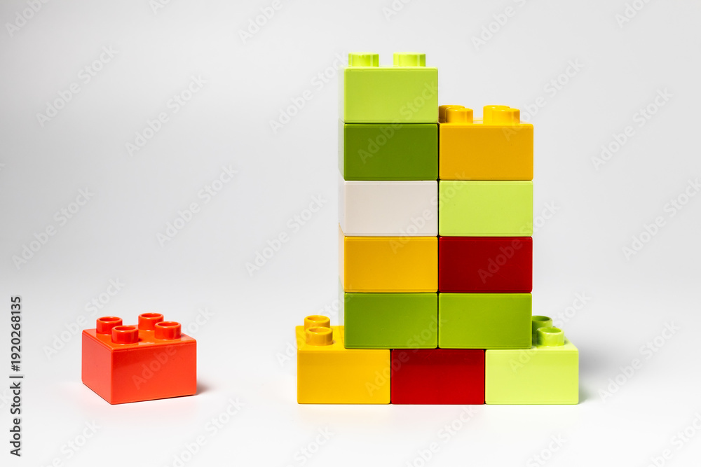 Fototapeta premium Colorful plastic building blocks stacked in a tower with a single red block placed separately on white background. Concept of teamwork, individuality, competition and business growth.
