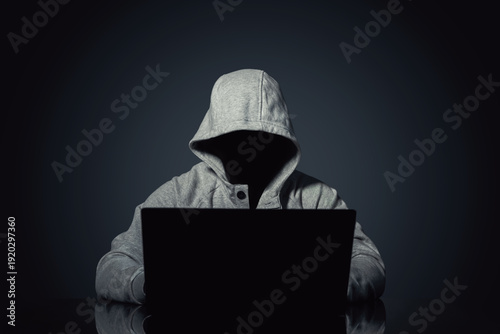 Faceless hooded figure using laptop on dark background, anonymous hacker concept, cybercrime, data breach, online security threat, internet blocking, privacy risk, digital attack scene at night
