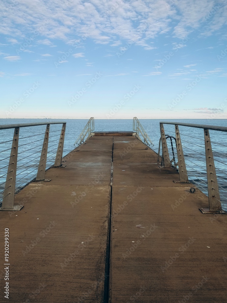 Obraz premium A long, narrow wooden pier stretches out toward a calm sea under a clear blue sky with soft, wispy clouds. The symmetrical perspective creates a sense of peaceful solitude and infinity.