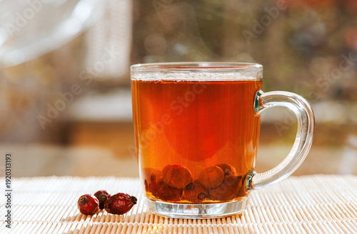 Transparent glass cup of herbal tea with rosehip berries glowing in warm sunlight on a natural bamboo mat.