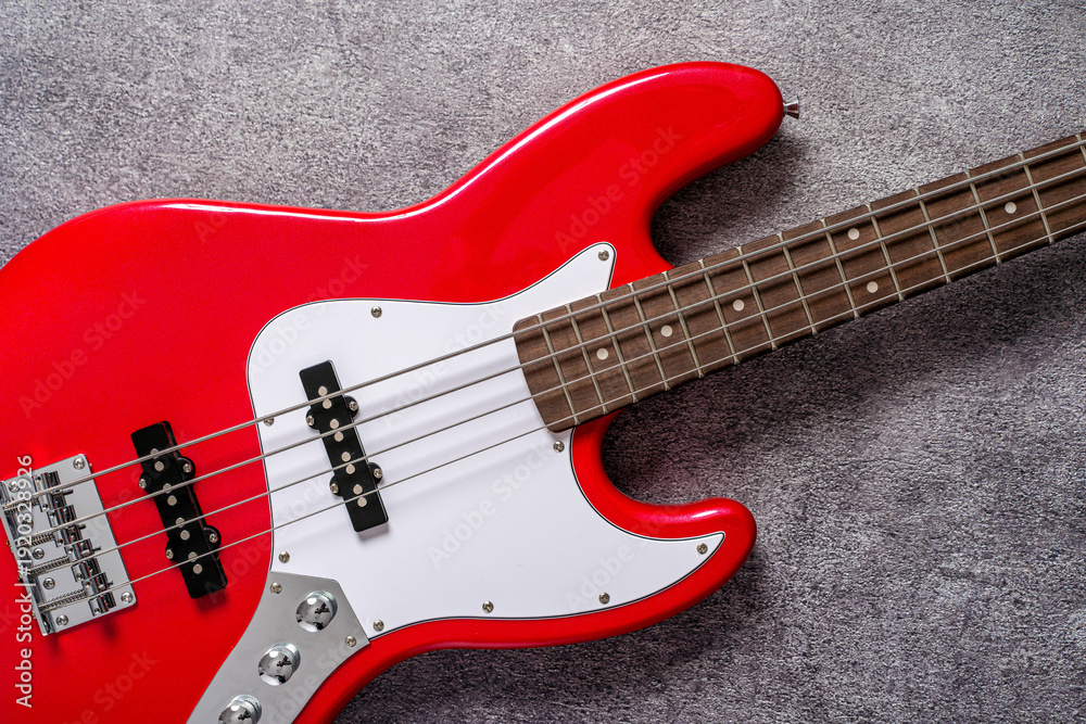 Fototapeta premium Detail of a red electric bass guitar with visible fretboard and headstock on neutral textured background. Music instrument concept, rock and funk sound, studio close up.