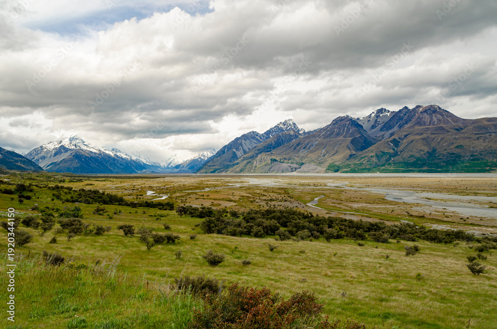 Fototapeta premium Rangitata River, with Mount Cook and surrounding mountains in the background. The scene includes a lush green foreground with shrubs and rolling hills, along with cloudy skies.