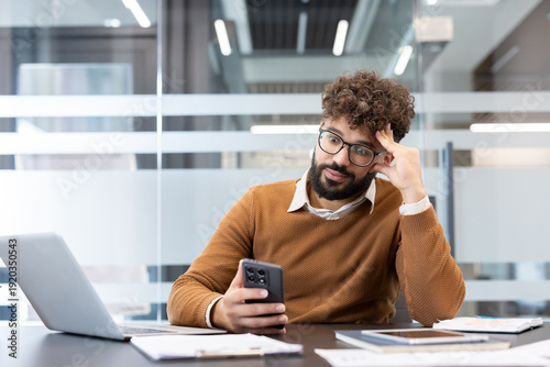 Photos Young man with curly hair and beard sits at an office desk holding a smartphone,