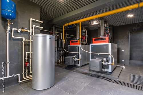 Two large boilers sit side by side in a utility room. A metal storage tank stands nearby. Pipes run along the walls and connect the equipment. The room is well-lit and features gray tiles