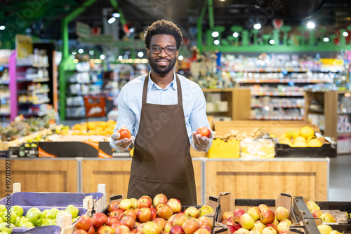 Slika na platnu Smiling male grocer wearing an apron and gloves, standing in a supermarket produ