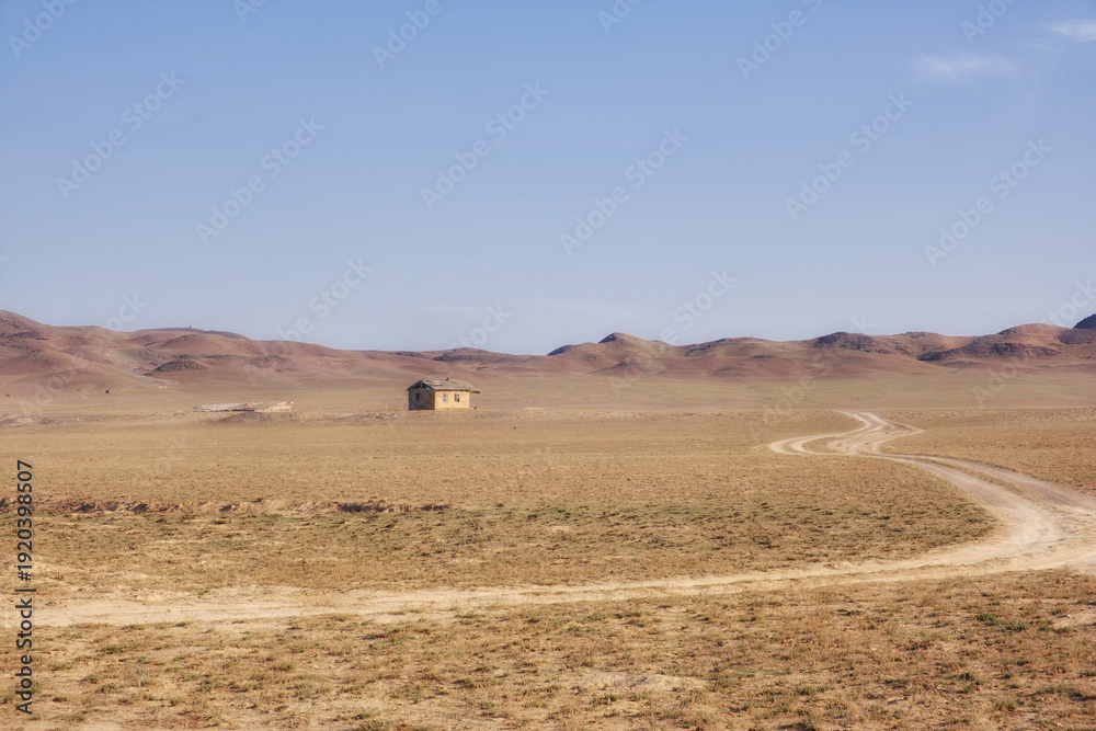 Obraz premium A lonely abandoned village in the endless steppe of Kazakhstan. A desert landscape with a winding dirt road leading to an old building set against a backdrop of low hills. 