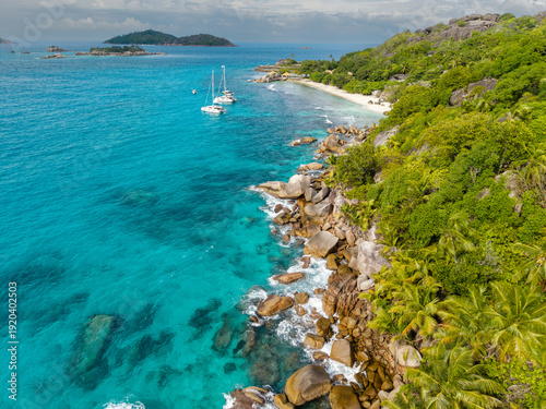 Aerial view of turquoise waters meet the rugged, green coastline near a secluded beach with boats offshore, Felicite, Seychelles.