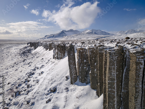 Aerial view of striking basalt columns stand sentinel against a backdrop of snow-dusted mountains under a sky brushed with wispy clouds, Raudhamelur, Eyja- og Miklaholtshreppur, Iceland.