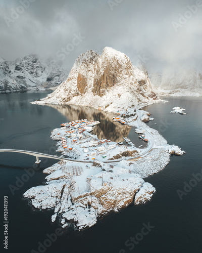 Aerial view of snow-blanketed Hamnoy, where red-roofed buildings nestle amidst the stark, rocky landscape against the steely sea, Nordland, Norway.