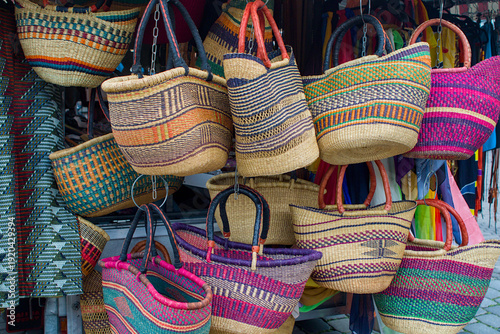Colorful woven baskets hang at a vibrant market stall. Striped patterns and curved handles create a lively display of craft. Natural fibers and bold dyes reflect rich artisan tradition.