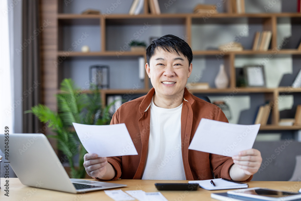 Fototapeta premium Asian man sitting at desk in home office, holding papers and looking at camera, actively managing personal finances and household budget with a laptop, feeling successful
