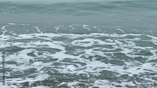 Waves roll onto the shore at the beach, creating a foam pattern on the wet sand as clouds cover the sky in the late afternoon light.