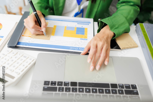 Fotografie A woman in a green jacket is talking on a tablet while sitting at a desk
