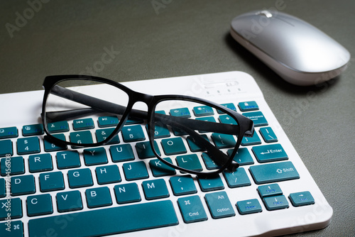 Black reading glasses resting on a wireless keyboard with teal keys next to a silver mouse on a dark desk mat. Perfect for technology, business and remote work concepts.