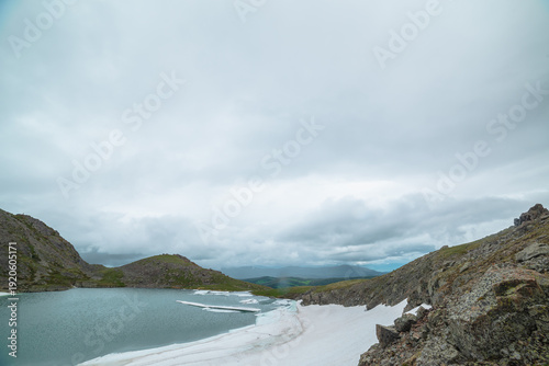 Alpine lake among snows in hanging valley with view to green hills far away u...