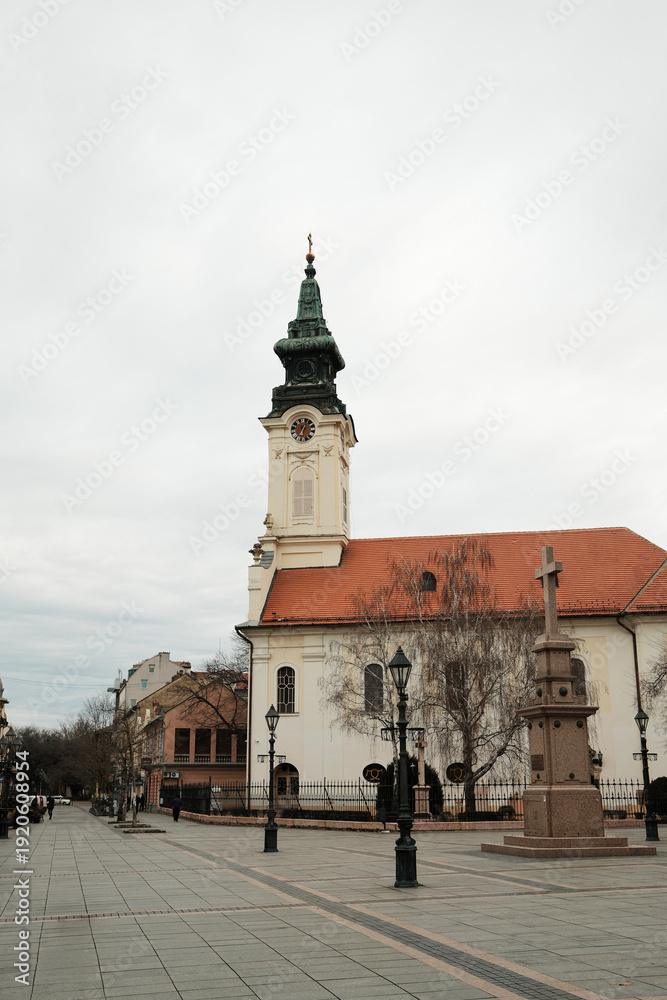 Obraz premium Church with clock tower and stone cross monument on open square in Sombor, Serbia. European religious heritage and city landmark concept.