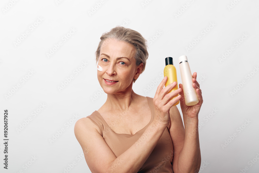 Fototapeta premium Mature woman with natural skin holds two cosmetic bottles, smiling gently. Skincare, beauty, and wellness concept focused on aging care and natural facial treatment on white background.