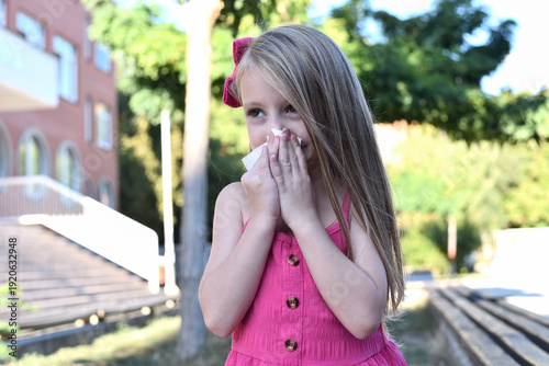 A little blonde girl sneezing into a tissue outdoors on a sunny day. Child suffering from cold or seasonal allergies. Health care and hygiene concept with copy space.