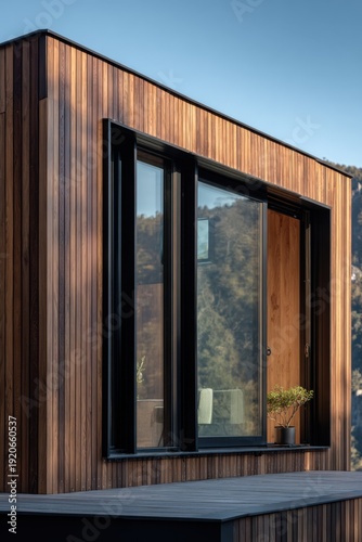 The wooden house features expansive windows that beautifully showcase the serene natural landscape in the background, with majestic mountains standing tall, all illuminated by bright daylight