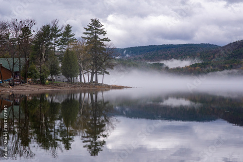 Typical lakeside cottage landscape on Lake Champlain characterized by a calm body of water reflecting the surrounding forest and enhanced by morning mist