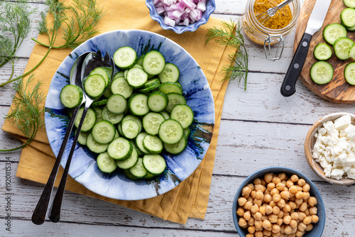 A bowl of sliced mini cucumbers with ingredients around for making a salad. 