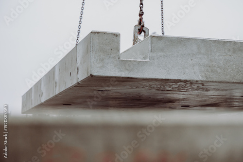 Large prefabricated concrete slab suspended by chains during crane operation. Heavy precast element positioned at an urban construction site.