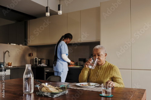 Senior woman drinking water while caregiver prepares meal in kitchen
