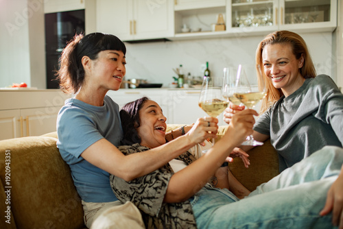 Three women friends toasting white wine on sofa at home