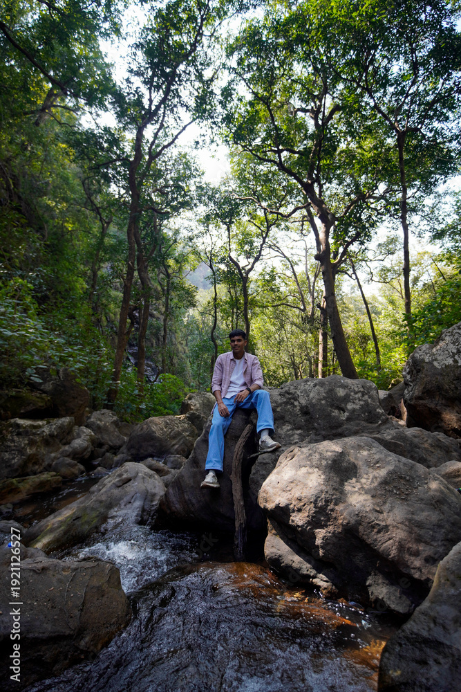 Obraz premium Young man relax on rock in lush green forest near flowing river stream