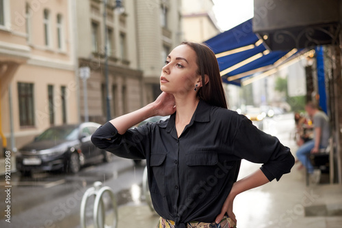 A woman in a black shirt and colored trousers stands on a wet sidewalk. The city sprawls behind her, with cars and buildings visible in the background.