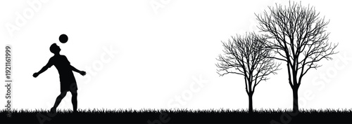 Silhouette of a person playing soccer with a ball in front of two bare trees on a grassy field
